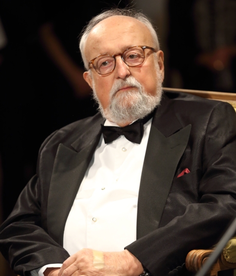 Krzysztof Penderecki siedzi na scenie w czarnym smokingu z muszką. Kompozytor spogląda przed siebie, a w tle widoczni są uczestnicy wydarzenia.