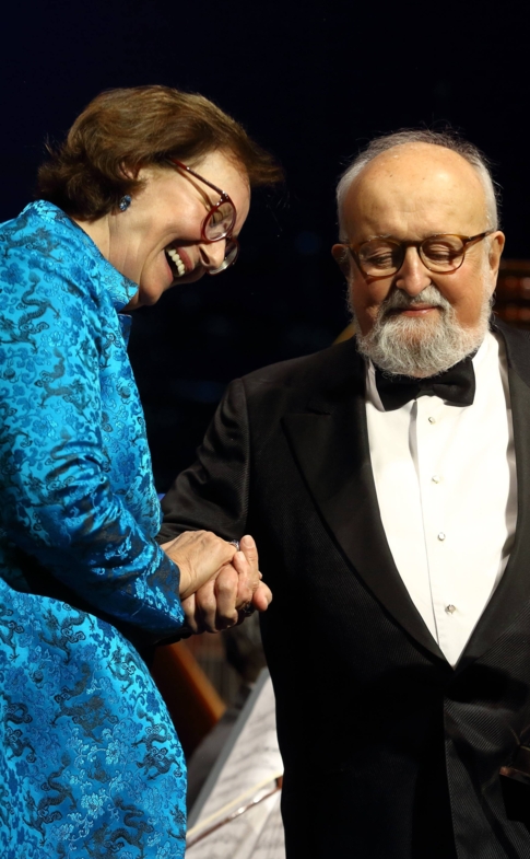 Krzysztof Penderecki odbiera statuetkę od kobiety ubranej w niebieską suknię podczas uroczystości na scenie. W tle siedzą muzycy orkiestry.
