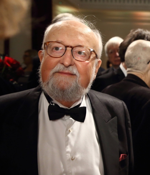 Krzysztof Penderecki i jego żona Elżbieta rozmawiają z uczestnikami podczas festiwalowego przyjęcia. Kompozytor ubrany jest w smoking z muszką, a w tle widać gości zgromadzonych w sali koncertowej.