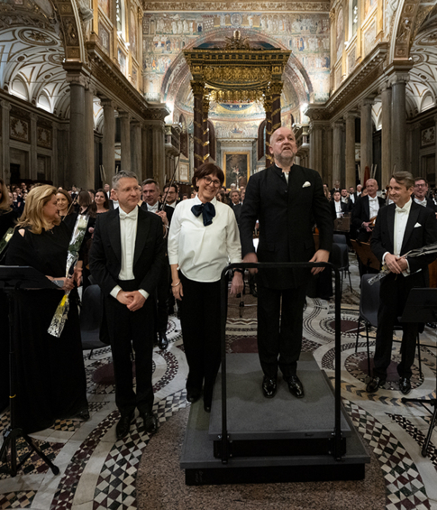 Dyrygent i soliści stoją przed orkiestrą po zakończeniu koncertu Credo w Bazylice Santa Maria Maggiore; część wykonawców trzyma kwiaty.
