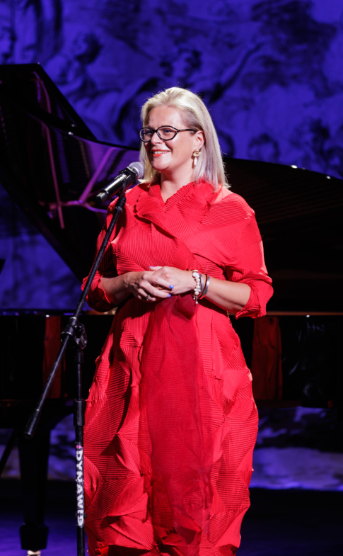 Woman in a red dress speaking into a microphone on stage, with a grand piano in the background.