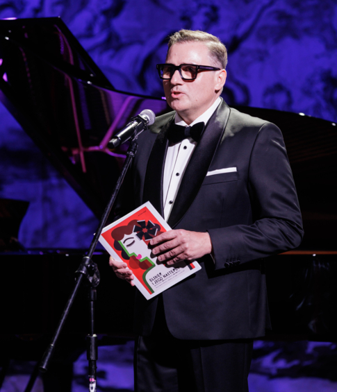 Man in a tuxedo speaking into a microphone while holding the event programme.