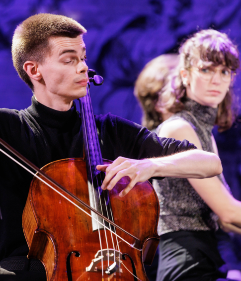 Close-up of a cellist performing, with the pianist visible in the background.