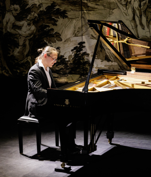 Pianist performing a recital at a Steinway & Sons concert grand piano against a decorative tapestry backdrop.