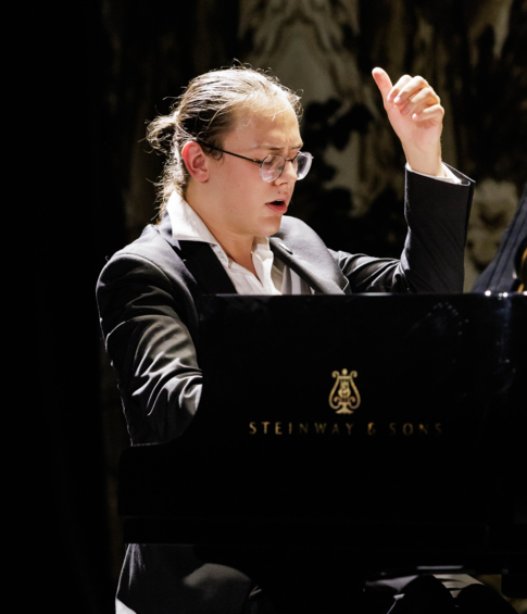 Pianist at a Steinway & Sons grand piano during an expressive moment of the recital, with one hand raised.