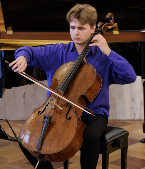 A cellist in a blue shirt performs during a chamber music concert, with a grand piano in the background.