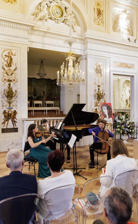 A violinist, pianist and cellist perform for an audience in an ornate historic concert hall.