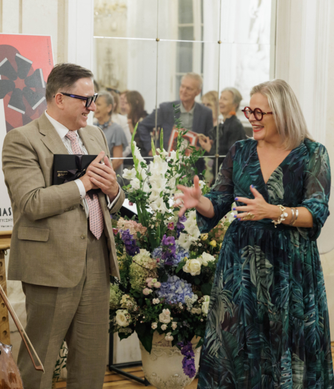 A cellist stands with his instrument beside a formally dressed woman and man at a music event, with flowers and a festival poster behind them.