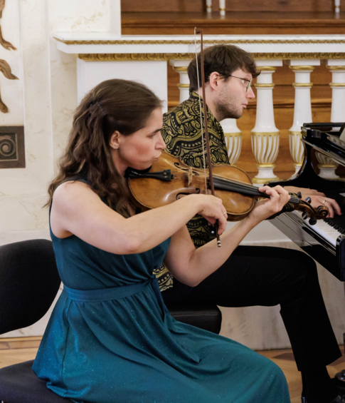 A piano trio—violinist, pianist and cellist—performs for an audience in a historic concert hall.