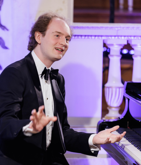 A pianist in a black tuxedo at a grand piano during a concert in a historic hall.
