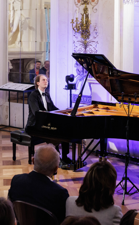 A pianist performs for an audience in an ornate concert hall illuminated with purple light.
