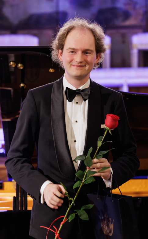 A smiling pianist in a tuxedo holds a red rose after the concert.