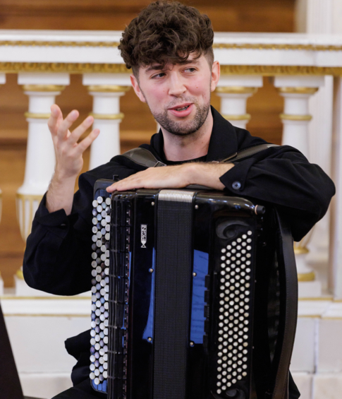 Akordeonista siedzący z instrumentem gestykuluje podczas wypowiedzi w trakcie wydarzenia.