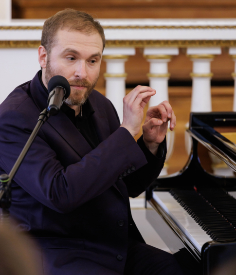 Pianista gestykuluje podczas wypowiedzi do mikrofonu przy fortepianie.