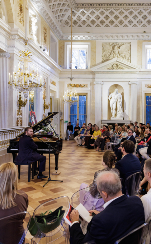 Pianista przy fortepianie przed liczną publicznością w bogato zdobionej zabytkowej sali koncertowej.