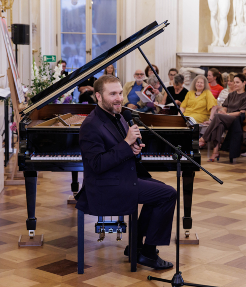 Pianista siedzi przy fortepianie i mówi do mikrofonu przed zgromadzoną publicznością.